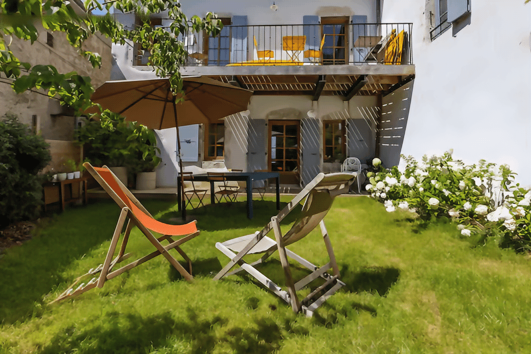 Sunny garden with deck chairs and umbrella, facing a charming house with a balcony.