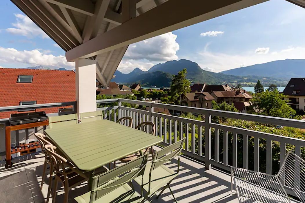 Balcony with table and chairs overlooking mountains and a lake under a sunny sky.