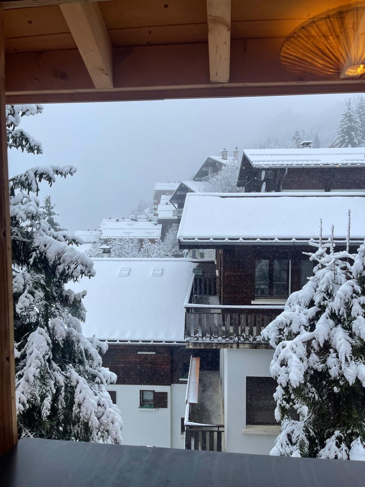 Snow-covered chalets and trees viewed from a wooden-framed window on a snowy day.